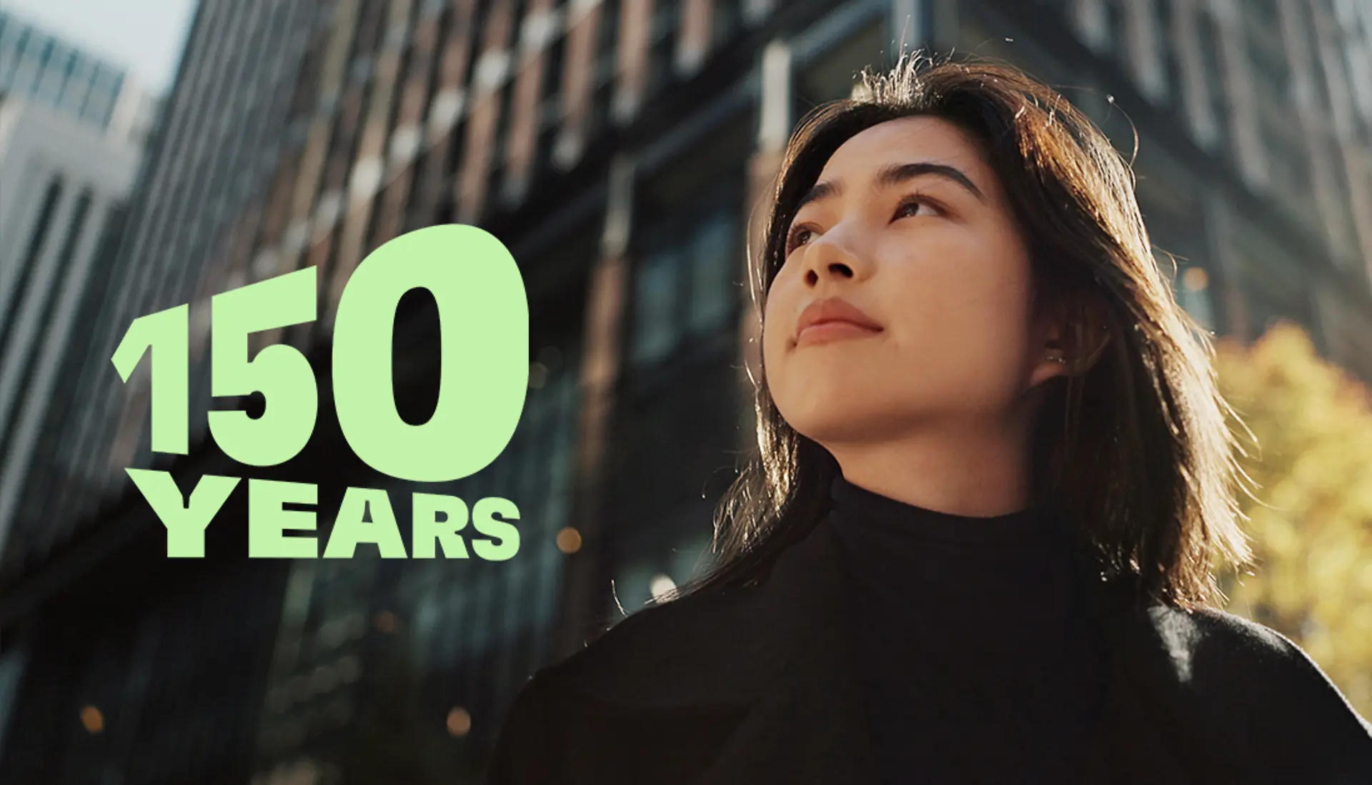
Green text reading ‘150 YEARS’ over a modern cityscape with tall glass buildings and autumn trees, symbolizing the 150th anniversary celebration.